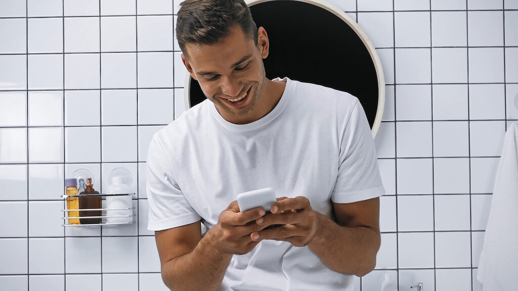 Man standing in bathroom smiling while texting on his phone.