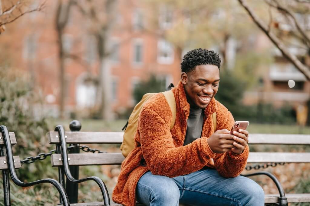 man sitting on bench, smiling while he is texting on his phone. 