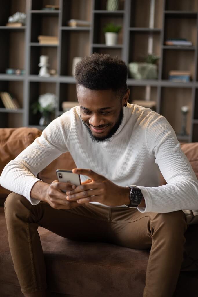 Man sitting on couch smiling while texting on his phone. 