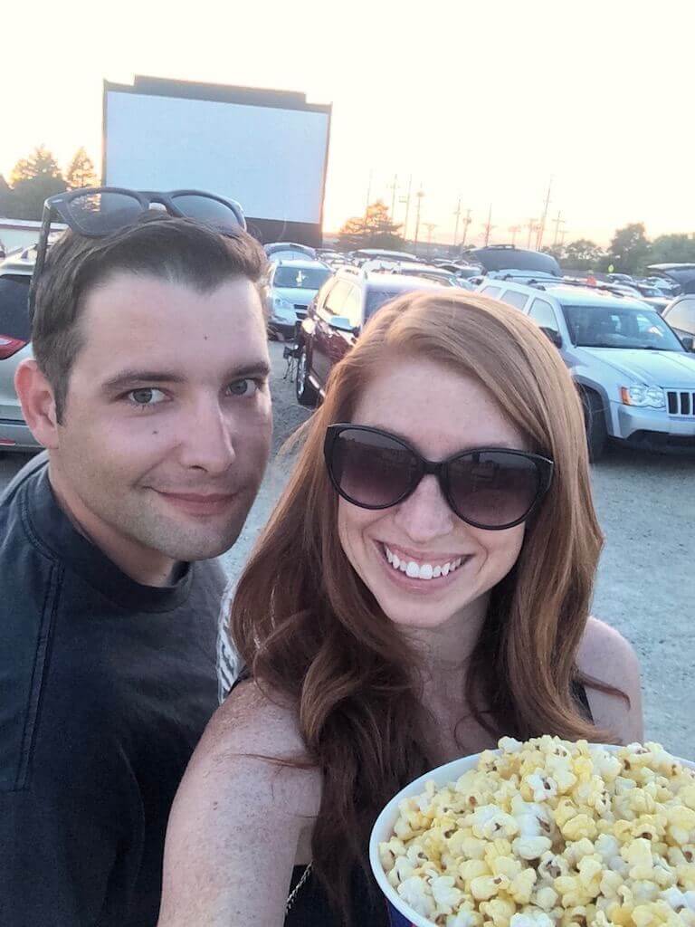 Couple on a date night, holding popcorn in front of a drive in movie theater screen. 