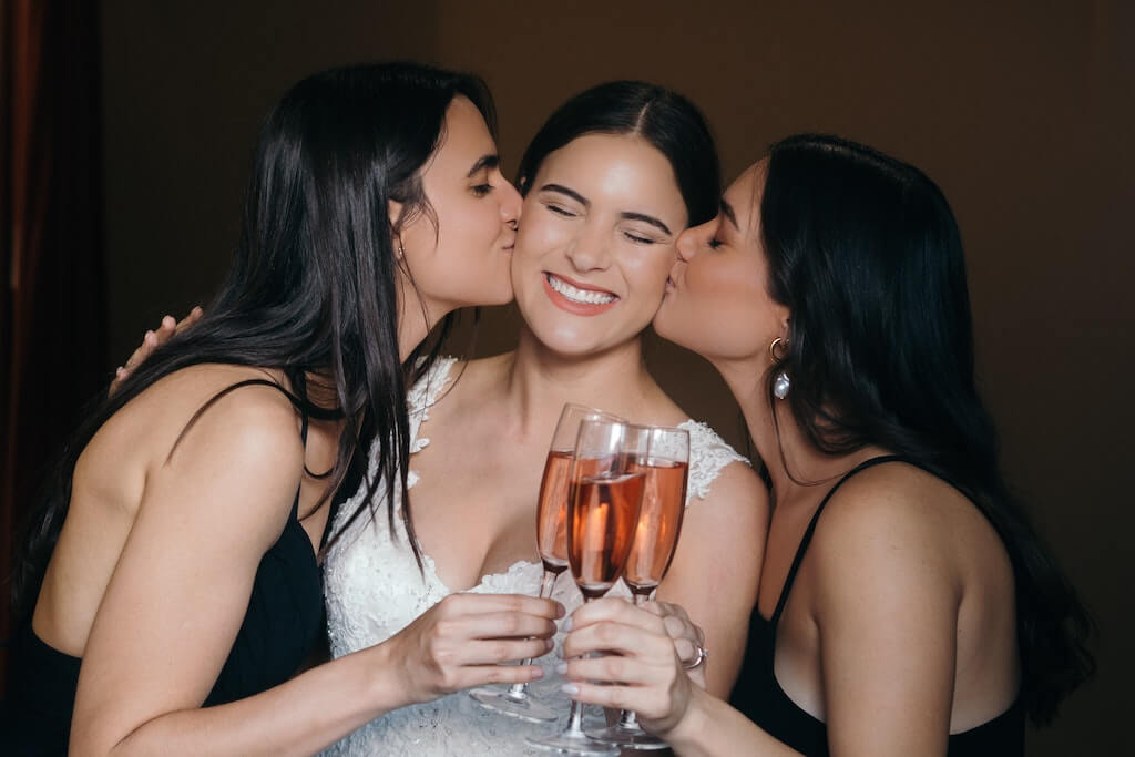 bridesmaids kissing brides cheek while holding wine glasses.