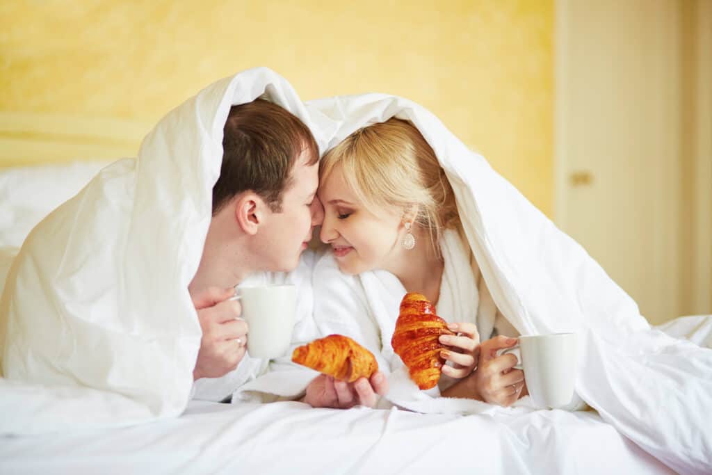 couple snuggling under covers and sitting coffee while eating a croissant. 