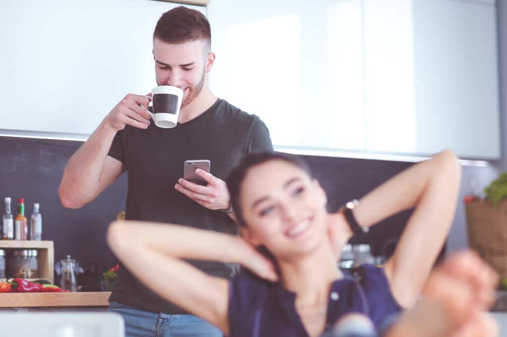 image shows a woman smiling and a man behind her looking at his phone smiling while drinking coffee.