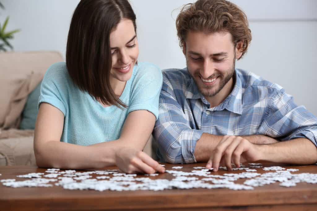 image shows a couple sitting at their dining room table, smiling and putting a jigsaw puzzle together.