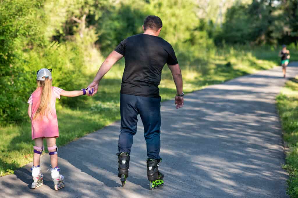 A dad holding his daughter's hand to support her while they both skate through the park on rollerblades.