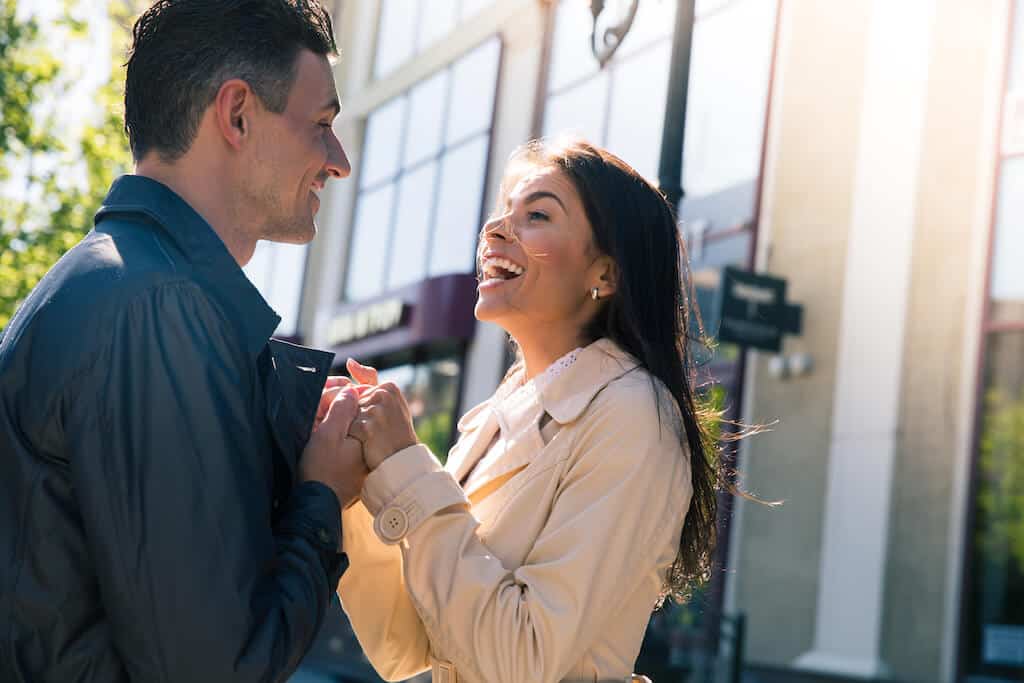 couple holding hands, facing each other and laughing outside. 