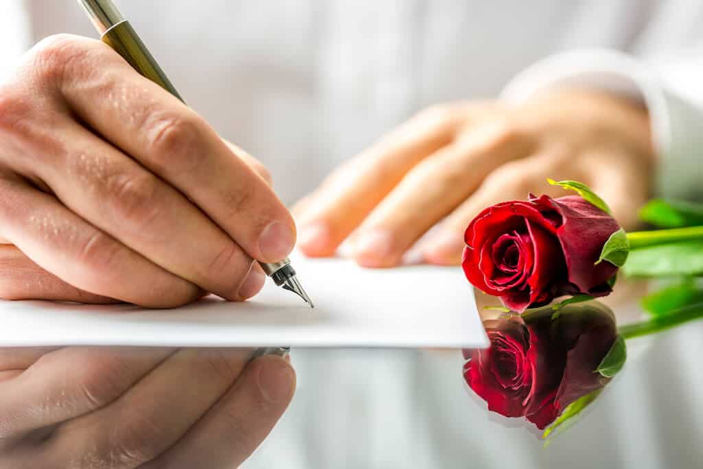man's hands writing a love letter with a rose next to it. 