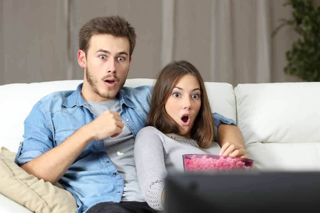 couple sitting on couch watching TV with shocked faces.